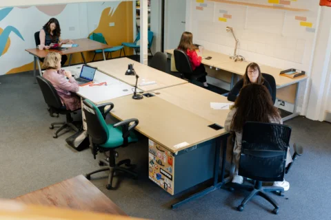 A photo of the Albany coworking space from above. Five people are sat working around a central table and two desks. The walls are covered with colourful murals.