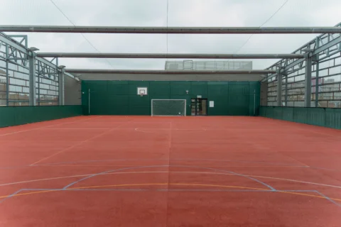 A photo of a ball court with red floor and green walls. The court is open to the elements and it is a grey, cloudy day.