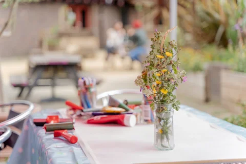 A photo of a table in the Albany garden, with a vase of dried flowers, paper, paints and paint rollers on it.
