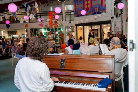A photo of the Meet Me choir performing in the Albany cafe. The view is from behind the pianist, looking over the piano to the choir and audience beyond.