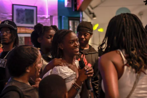 A group of young people gathered in a lively indoor space, with one girl in the centre smiling and holding a microphone.