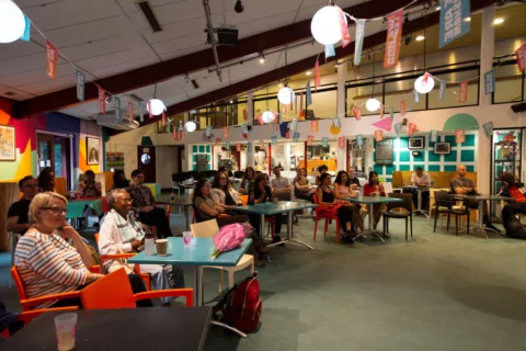 A diverse audience of many ages sat inside a colourful cafe space. All look very engaged and attentive.