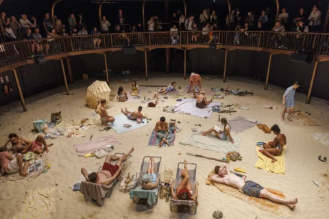 A view looking down at the Albany auditorium from the balcony. The floor is covered in sand to resemble a beach, with performers sunbathing on sun loungers and towels.