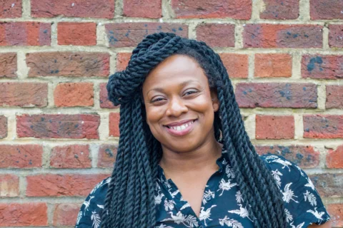 A photo of Vicki Amedume, a black woman with long braids, stood in front of a brick wall.