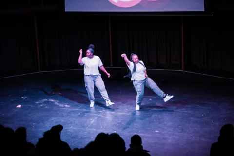 Two young dancers in a stage under spotlight. They are moving in sync and the audience is silhouetted in the foreground of the image.