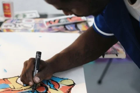A close up photo of a black man leaning over a table, drawing. He is using a black marker pen to outline an illustration of a face, coloured in with blue, orange and yellow.