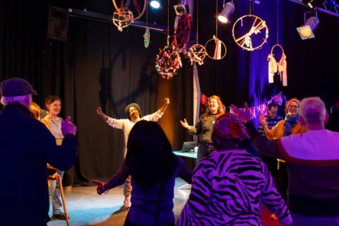 A group of people stand in a circle with their arms raised (they are doing a dance warm up). The room is surrounded with black curtains and there is purple lighting, and dream catchers hanging from the ceiling.