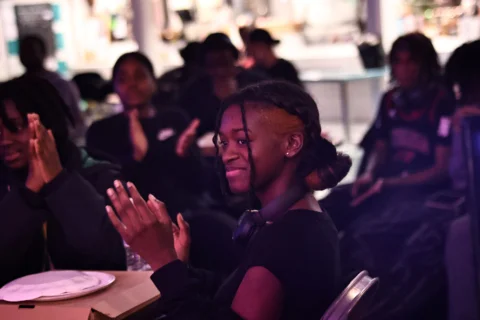 A photo of a young black girls sat in a crowd. She is wearing a black hoodie, looking off to one side and smiling and clapping.