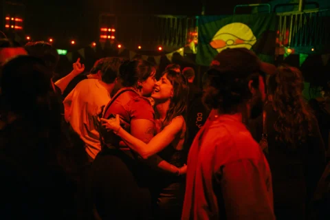 A photo of two women hugging in the middle of a crowded dance floor. They are bathed in red lighting.