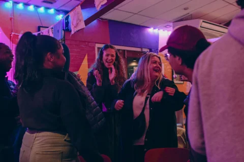 Photo of a group of people chatting and laughing in the Albany cafe. There is a colourful mural in the background and they are bathed in a purple light.