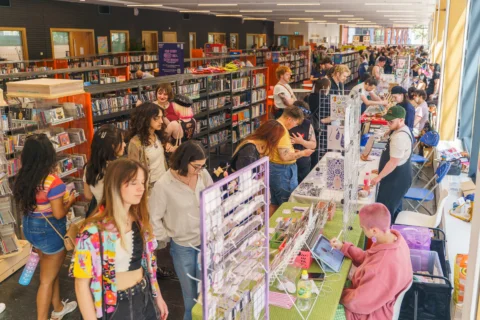 A row of craft stalls in the library at Deptford Lounge. The stalls are parallel to the bookshelves in the library and people are milling around, chatting to the stallholders.