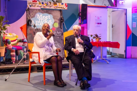 Elderly woman and man smiling, seated with microphones, against a colorful stage backdrop. Vibrant, inviting atmosphere with warm lighting.