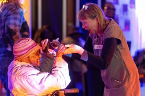 A woman is holding the hands of, and dancing, with another lady in a wheelchair. Both women are smiling enthusiastically at each other giving a joyful and excited.