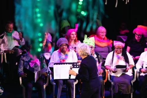 A vibrant choir lit by pink stage lights, featuring singers of various ages, performs under the guidance of an enthusiastic conductor.