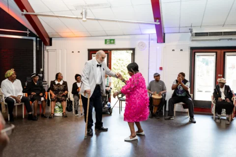 There are two older people, dressed up very smart in a suit and pink dress, they are holding hands and dancing in the middle of a bright room with large windows. There are several other people sat in chairs in a circle around them. These people are smiling, clapping and even one man playing a drum. The atmosphere is joyful and lively.