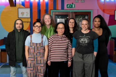 Seven people stand indoors in a row, posing for a group photo against a colorful mural background.