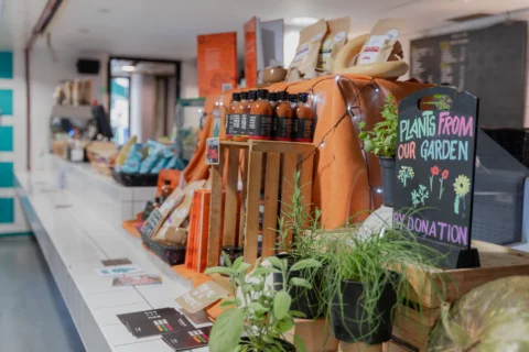 A photo of the counter in the Albany cafe. It has white tiles with snacks, chutneys and pots of herbs displayed on it. A small black board has 'Plants from the garden by donation' written on it in coloured chalk.