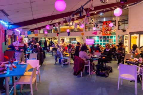 A photo of the Albany cafe during a Meet Me party. The tables are full of people chatting, there is bunting hanging from the ceiling and pink lighting.