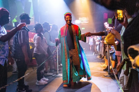 A photo of a young black man wearing bright colours on a catwalk. There is brightly coloured lighting and people on either side of the catwalk are cheering him on.