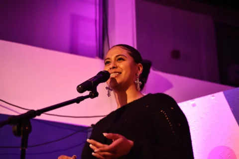 A young woman standing in front of a microphone on stage. The lights are a pink hue and the scene captures her focus she performs indoors, with a smiley demeanor.