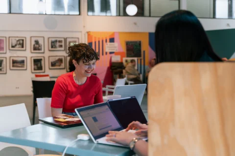 A photo of two people sat at a table on laptops. One has their back to the camera. The other is a white woman with a red top and curly hair, she is smiling.