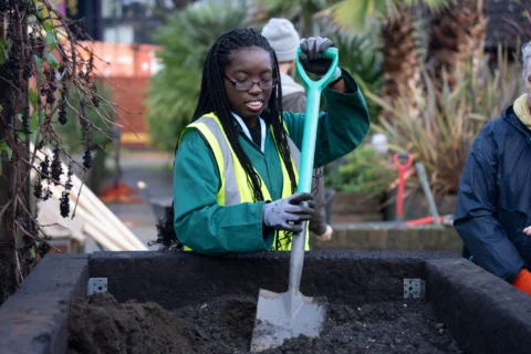 A young girl is holding a shovel in the action of planting a tree in a free standing planter outside. She is wearing a hi-vis vest and smiling.