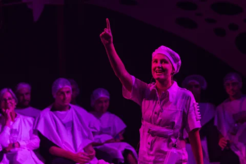 A performer in a white uniform and hairnet, illuminated by purple light, energetically gestures by pointing towards the sky on a stage. An attentive audience in similar attire watches.