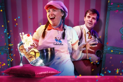 A photograph of two performers against a colourful pink stripy background. In the foreground is a white woman with plaits and wearing a pink hat and apron, holding an ice cream sundae which has been presented in a high heeled show. A white man peers out from behind her looking on.