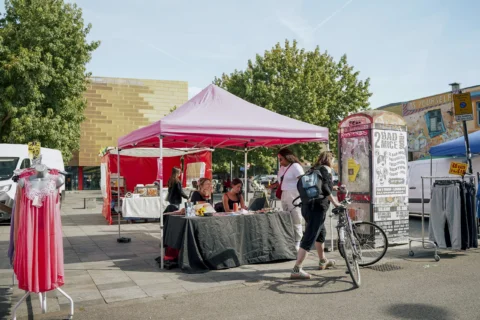 A photo of a stall on Deptford Market. It is a sunny day and the stall is under a bright pink gazebo. Someone holding a bike is stopped in front of the stall and there are trees and other stalls in the background.