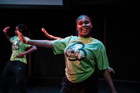 A young woman energetically dances on stage as part of an ensemble, all are wearing a green "Love2Dance" shirt. She's smiling, with arms outstretched, conveying joy and enthusiasm.
