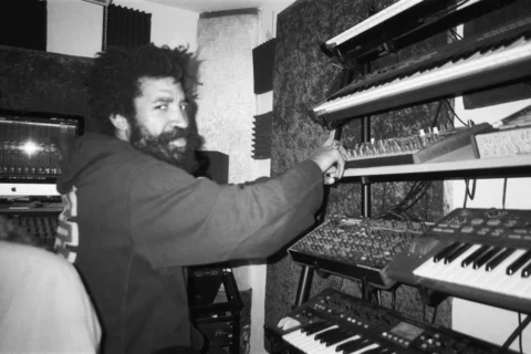 A black and white photo of musician Kwake Bass - a man with an afro, beard and wearing a hoodie, stood in front of a wall of keyboards and DJ decks.