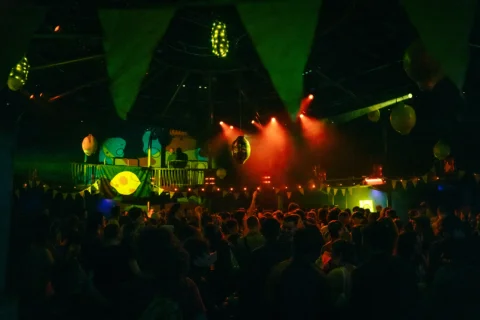 A photo of a club night in the Albany theatre. The auditorium is full of people dancing. It is dark but there are red and green beams of light and a yellow lemon is projected on the far wall.