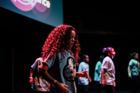 A young girl with long brown curly hair and wearing a blue t-shirt is dancing on stage. Behind her are a group of other children all wearing blue t-shirts.