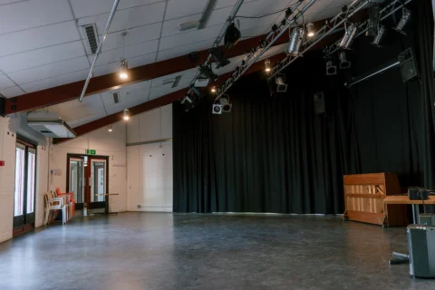 A photo of the Red Room at the Albany. It's an empty space with a lighting rig on the ceiling, and black curtains surrounding the walls. There is a piano in the corner.