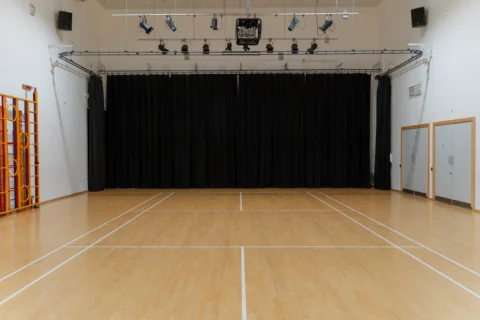 A photo of the Main Hall at Deptford Lounge. It looks like a school sports hall, with court lines on the floor, and gymnastics equipment along one wall. There is a lighting rig on the ceiling and a black curtain covering the back wall.