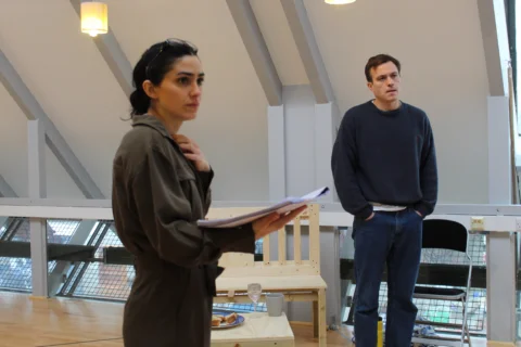 Cassie Bradley and Barney White in rehearsal for One Day When We Were Young. A woman wearing a grey boiler suit stands with one hand holding a script and the other on her chest. Behind her is a man wearing jeans and a black jumper with his hands in his pockets.