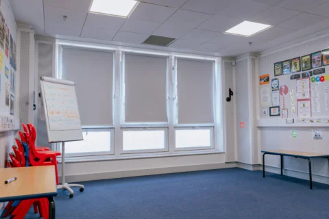 A photo of the music room at Deptford Lounge. It is a room with white walls and a blue carpeted floor. One wall has floor to ceiling windows with the blinds pulled most of the way down. In the space is a flipchart, two tables and a stack of orange and black chairs.