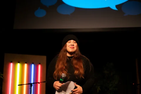 A person in a black outfit and long brown hair wearing a beanie holds a microphone on stage with a colourful neon light display behind them, conveying a bright and lively atmosphere.