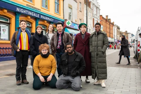 A diverse group of eight people, dressed warmly, gather on a lively Deptford High Street." The mood is cheerful.