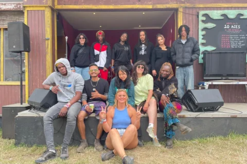 A diverse group of young people pose happily on a wooden stage outdoors. Some sit on the edge while others stand in a line behind them. The mood is relaxed and joyful.