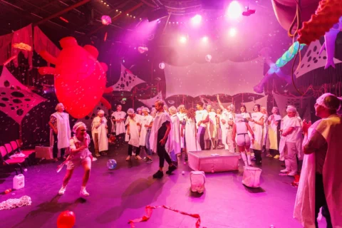 A lively, whimsical stage scene features people in white costumes and shower caps, brightly lit in pink. The backdrop is filled with colourful, abstract decorations.