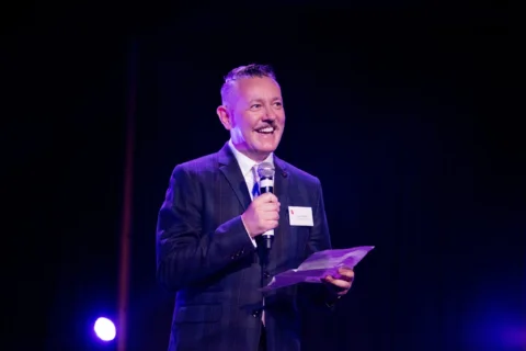 A man in a suit is smiling and holding a microphone on a dark stage. He is uplift in pink and blue, the mood is joyful and celebratory.