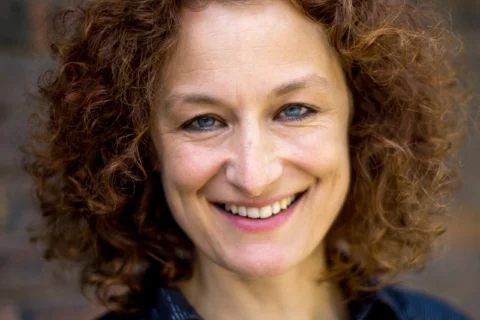 A woman with curly red hair and a friendly smile is wearing a black shirt. She stands against a blurred brick wall, conveying warmth and approachability.