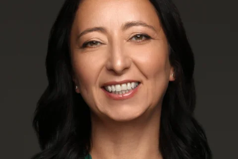 Smiling woman with shoulder-length black hair, wearing a teal top and a simple necklace, against a plain dark background. She conveys warmth and confidence.