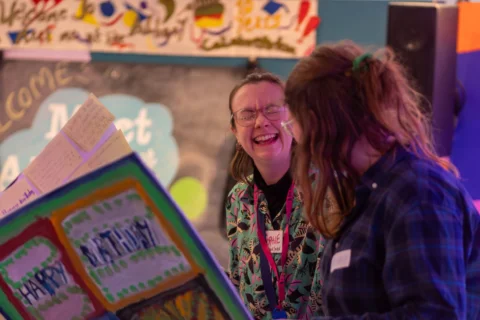 Two people laughing while looking at a comically oversized, handmade, colourful card with "Happy Birthday" on it. A festive, joyful atmosphere with a decorated backdrop.