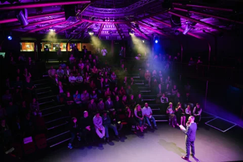 The image shows a theatre audience watching a sole presenter on stage. The spotlights are pink and vibrant. The speaker has their back to the audience and is wearing a suit, holding notes and speaking into a microphone.