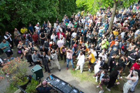 A lively outdoor party with a diverse crowd of people dancing and socialising. A DJ booth is visible, surrounded by green trees. Warm, joyful atmosphere.