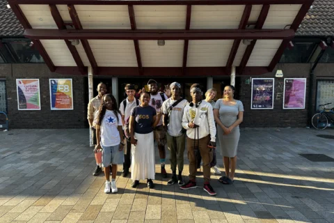A group of young people stand smiling outside the Albany front doors. The sun is shining bright, creating a warm and friendly atmosphere.
