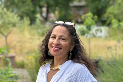 Woman in a white blouse smiles warmly in a lush garden setting. She wears sunglasses on her head and gold jewellery, creating a serene and joyful tone.