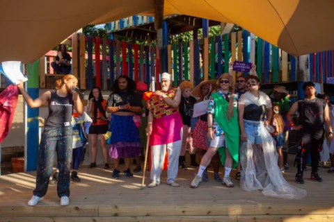 A lively group on a colourful stage celebrates, dressed in colourful, eclectic outfits. A person in holds a mic, creating an energetic, festive atmosphere.